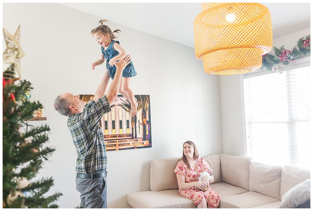 In a living room, a mom holds a newborn baby and smiles while a dad tosses big sister in the air.