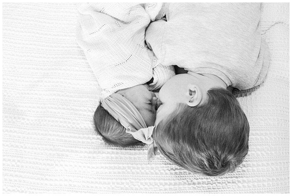 A black and white photo of a toddler and his newborn sister laying facing each other nose to nose.