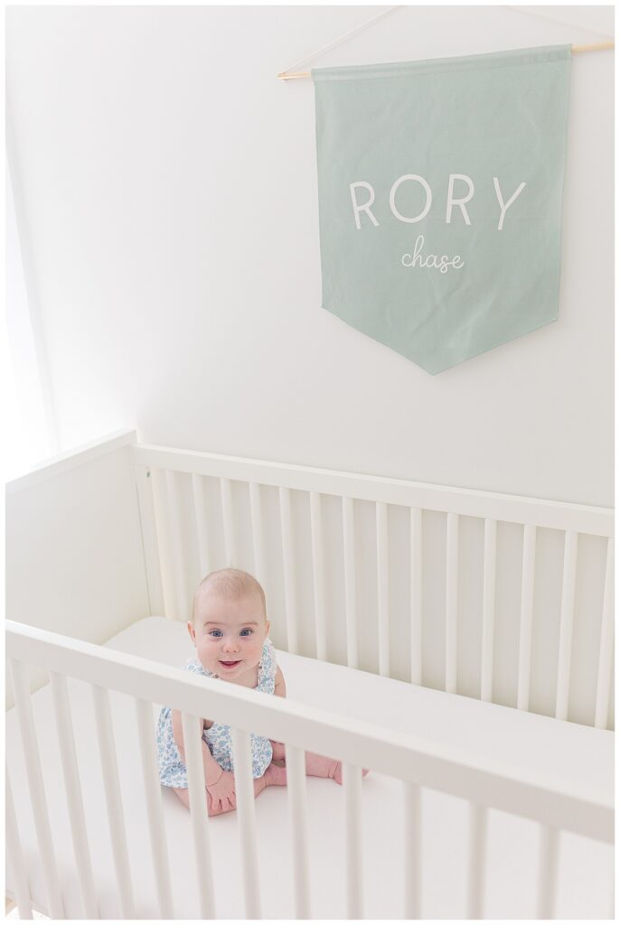 A 6 month old baby sits and smiles in her crib during a lifestyle newborn session with an older baby in Richmond, Virginia.