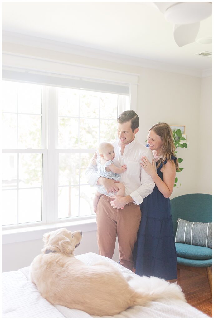 A mom and dad snuggle their daughter by a window while their family dog looks on during a lifestyle newborn session with an older baby in Richmond, Virginia.