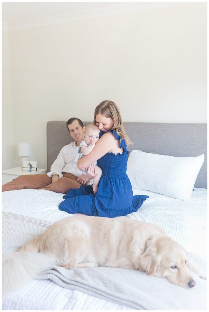 A mom snuggles her daughter while dad smiles at them and the dog rests at the foot of the bed during a lifestyle newborn session with an older baby in Richmond, Virginia.