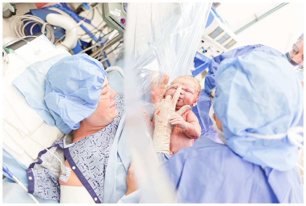 A new mom looks through a clear drape at her newborn baby as the doctor touches baby's nose after a c-section.